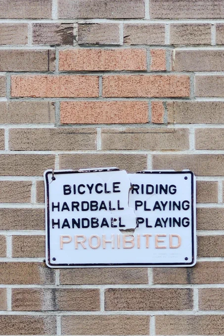 Cracked sign hanging on brick wall with text:
BICYCLE RIDING
HARDBALL PLAYING
HANDBALL PLAYING
PROHIBITED