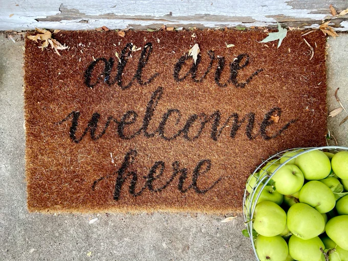 Thanks to LeeAnn Cline on Unsplash for the photograph of this welcome mat which reads All Are Welcome Here, with autumn leaves and a basket of crisp green apples!