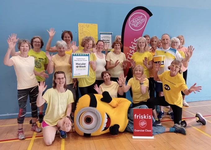 Group photo of the Friskis and Svettis Aberdeen fitness class celebrating their fundraising efforts. A person in a Minion outfit lies across the front, with everyone holding their hands in the air, smiling at the camera. Fitness leader Rosey is in the front row, far right, wearing yellow glasses.