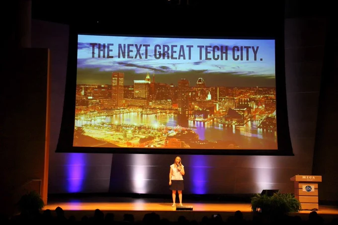 Woman in white shirt and black skirt speaks on brown stage in front of project with Baltimore skyline