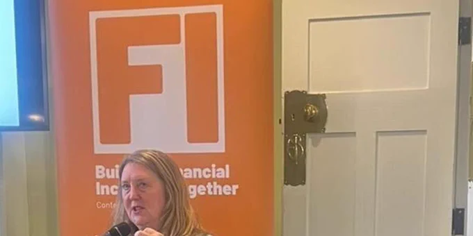 Our CEO, Helen Milner OBE, sitting on a panel at the Fair4All Finance Financial Inclusion Conference in London. Helen has blonde hair and is wearing a brightly coloured printed dress. She is holding a microphone. There is a banner in the background. 