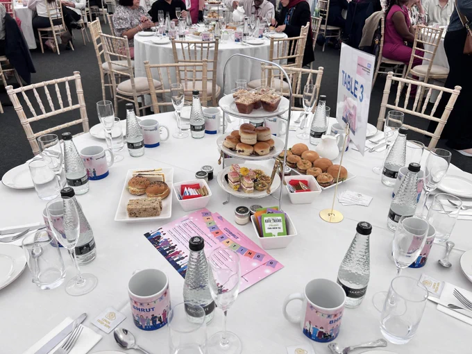 Image of the afternoon tea table. There is a plate tower with cakes, scones and sandwiches on it. There are champagnes glasses and mugs for tea. There is a menu on the table