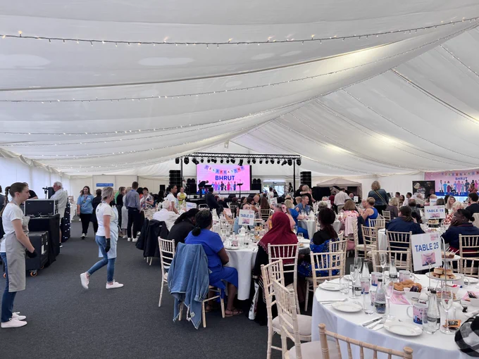 The Long Service Awards in the marquee. People are sitting at multiple tables in front of a stage. There is afternoon tea treats on the tables and everyone is chatting