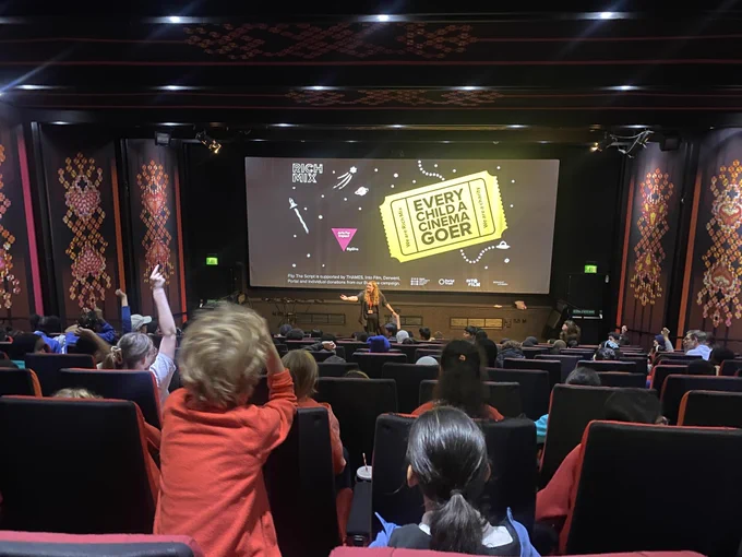 A person stands in front of a cinema screen with a slide reading 'Every Child A Cinema Goer' on it, as children sit in cinema seats with their hands raised in the air, answering questions.