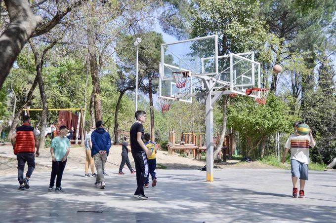 A group of teenage boys wearing casual spring attire play basketball in a park in Tirana.
