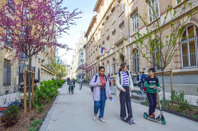Three children walk and scoot along a school street lined with plants and trees in Paris.