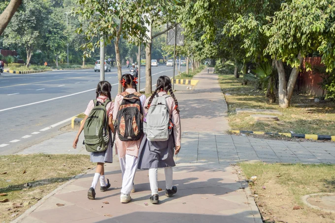 A group of three girls wearing school uniforms walk arm-in-arm along a footpath in Delhi.