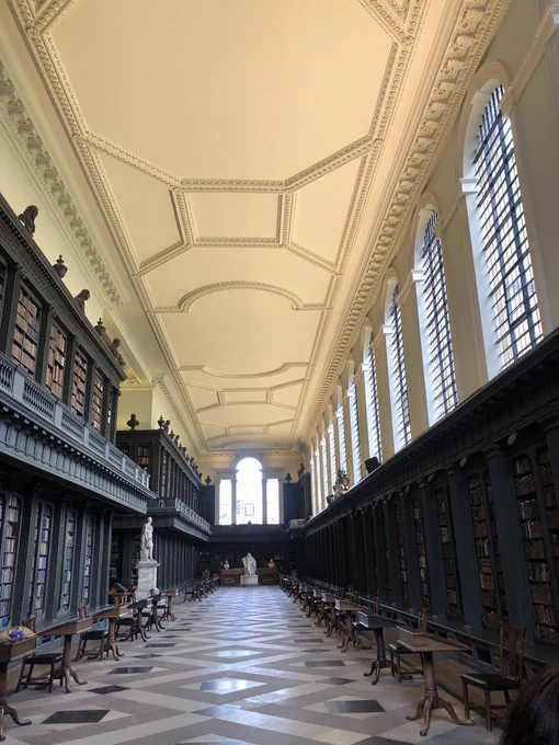 The main reading room of All Souls College Library, including two white statues, and two rows of sloping wooden desks and chairs. 