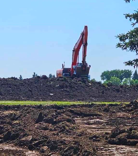 A red excavator sits in the middle of the image amidst trees in the background. There's a row of dirt in front of the excavator, a row of grass in front of that and a large stretch of overturned earth in the foreground.