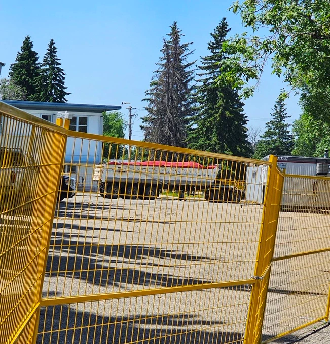 Behind a yellow construction fence sits two small buildings standing to either side of a trailer covered in a red cloth. There's several trucks to the left of the image in front of one of the buildings.