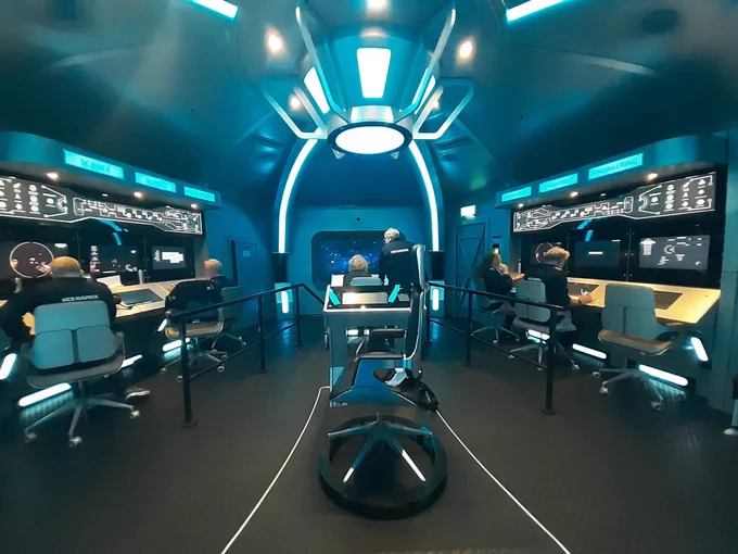The bridge of a spaceship with captain’s chair in the middle, helm, main screen, and three officer stations on either side. There’s half a dozen people in blue jumpsuits at the various stations and there is a lot of bluish lighting.