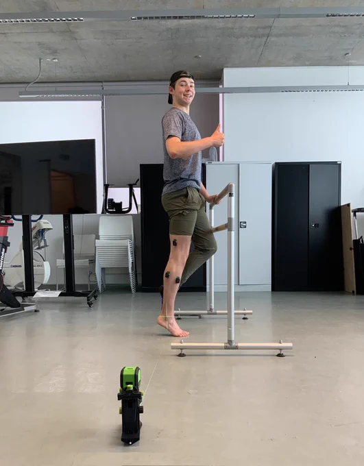 This is a picture of a young man wearing a baseball cap backwards with a thumbs up with EMG sensors on one leg in a relieve. There is also a laser aimed at his lateral maleolus.