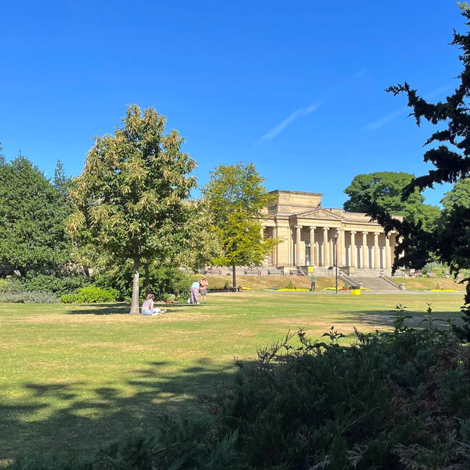 Weston Park on a bright sunny day under a blue sky. A person is sitting under a tree and the museum building is seen in the background.