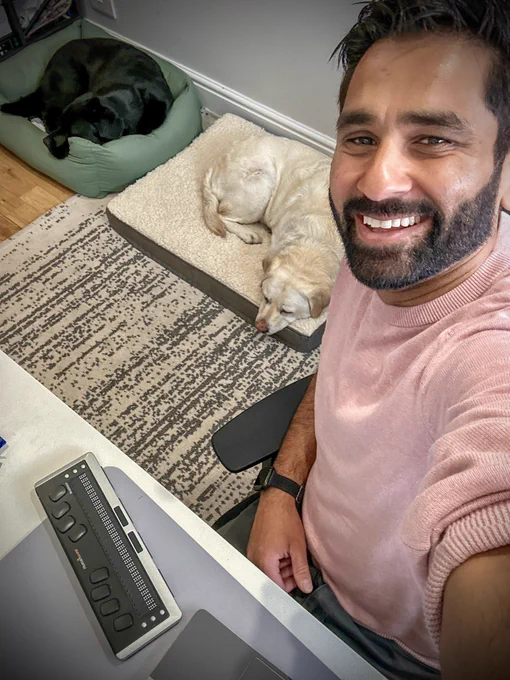 A selfie of Amit in his office.  He is wearing a pink jumper and smiling, you see kika sleeping on her bed stretched out and quark is snuggled in his.