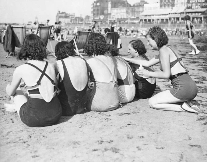 A black and white photo of a group a women at the beach. On the far right, a woman applies sun cream to her friends' backs on Margate Main Sands, 1934. Image credit: © Hulton-Deutsch Collection/CORBIS/Corbis via Getty Images