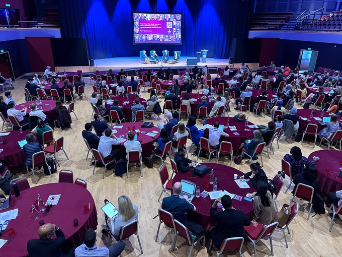 A birds eye view of a conference hall, with round tables with red table cloths, with many people sitting around the tables. At the front of the hall, is a large stage, with four green chairs, with the Papers Podcast team sitting. There is a large screen behind the chairs with a promotional graphic for the podcast. 