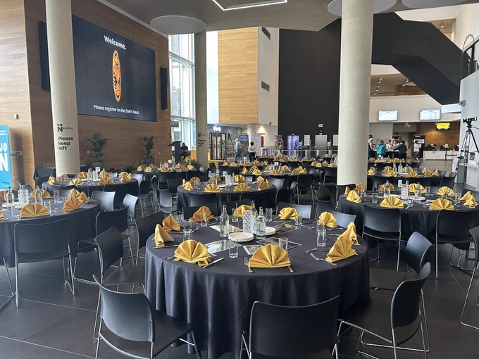 Image shows the Learning Hub at the University of Northampton which is set up for the People Awards 2024. There are round tables and chairs. The tables have black tablecloths and gold napkins. The tables are set with cutlery and glasses. In the background are some people registering those who are attending.