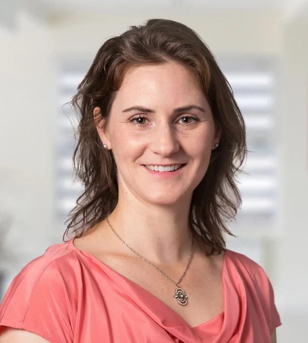 Portrait of a smiling individual with shoulder-length brown hair wearing a light pink blouse and a delicate necklace. The background is softly blurred with hints of a bright, window-lit room.