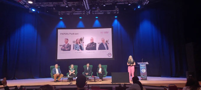 Photo of the podcast panel sitting on chairs on a stage 