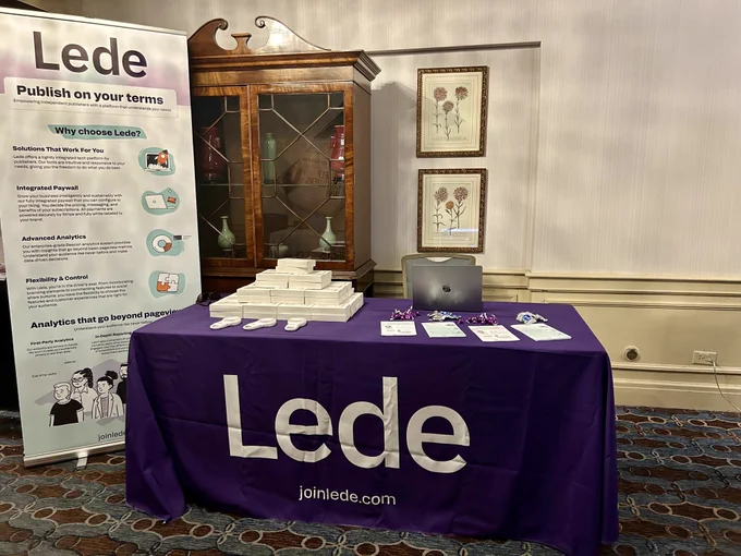 Lede's vendor table at the AAN conference is covered in a bright purple tablecloth that reads "Lede." On the left side of the table is our swag - a white handheld mini fan. Three fans are out of their boxes and behind those samples is a pyramid of fan boxes. On the right side of the table is an open laptop, some handouts, and some purple and silver candy. To the left of the table is a popup banner with info about Lede and some whimsical graphics, including a spaceship launching out of a computer. 