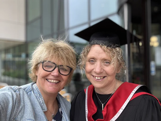 2 women smiling selfie , one a graduate in gown and hood.