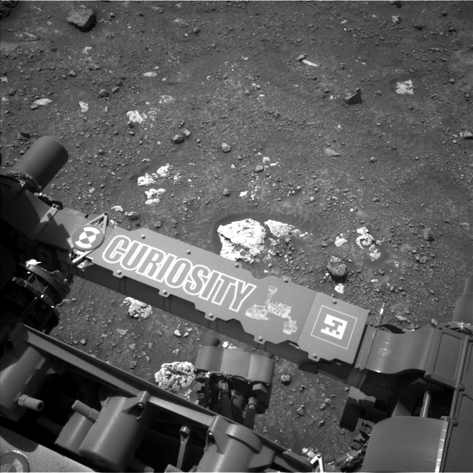 A black and white photo shows the nameplate for the Curiosity rover with a rough sandy Martian surface in the background. The rover's nameplate is diagonal in the image, stretching from the center left to the lower bottom right. The surface of Mars is sandy with dozens of small rocks and some larger white ones lining the area.