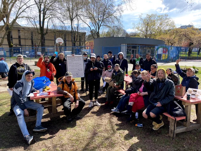 Twenty members of EIDGE meet in Oppenheimer Park. They smile at the camera while sitting on benches and standing around a big white pad. 