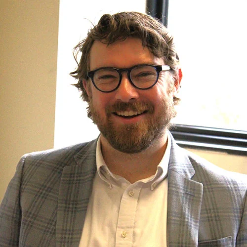 Photo of Noah Chauvin, a man wearing a white shirt with a checkered suit coat and glasses. 