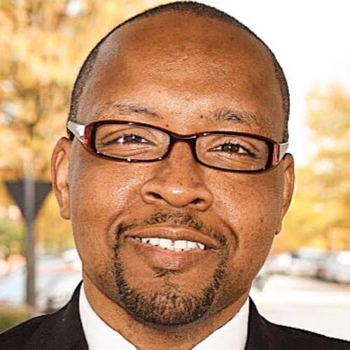 Photo of T.J. Peterkin, a man with a white shirt and black suit jacket. He is smiling and is wearing glasses. 