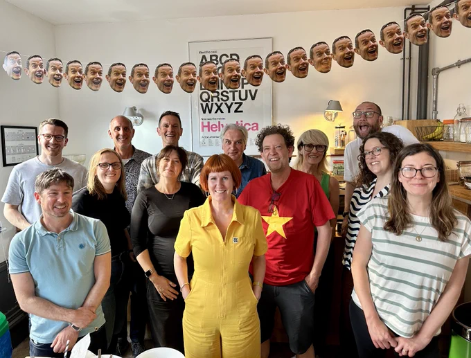 A group of 13 people are smiling at the camera in an indoor setting. One person in the front is wearing a bright yellow outfit, and another person is wearing a red shirt with a yellow star. The background features alphabet posters and a garland of printed faces.