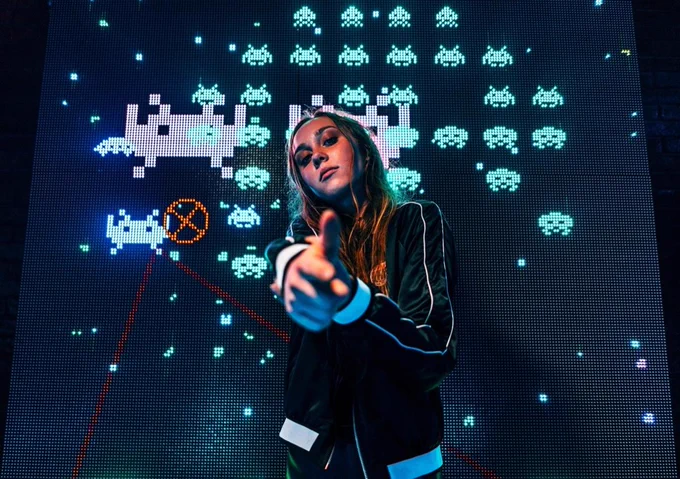 Chica apuntándote con un dedo como si fuera una pistola y con un fondo del video juego de invasores del espacio detrás.
