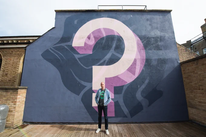 A person stands in front of a large mural painted on an exterior brick wall. The mural depicts a prominent, multicoloured question mark on a dark blue background. The person is wearing a green shirt, blue jacket, and dark pants, with hands in their pockets.