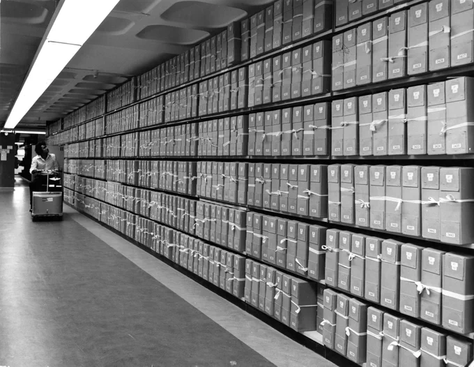 A black and white photograph from the late 1970s of a person in the stacks of the then Public Record Office. This photograph is from an album of photographs showing the construction of, and transfer of records to the Public Record Office, Kew.