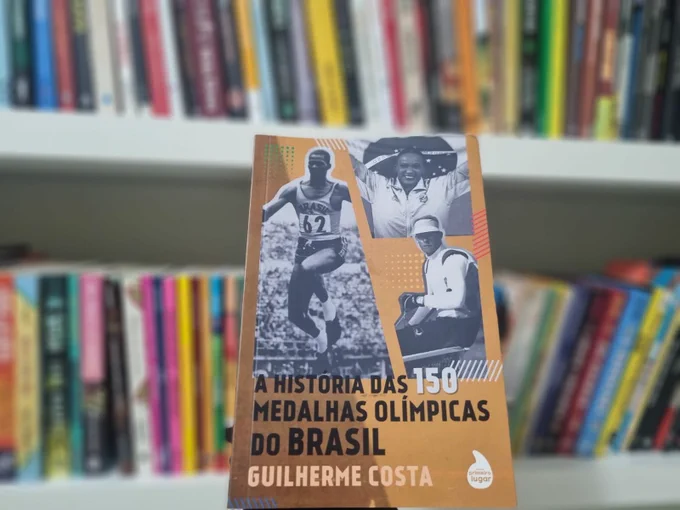 Livro em primeiro plano com o título A História das 150 Medalhas Olímpicas do Brasil, Guilherme Costa. Com vários livros atrás em segundo plano.
