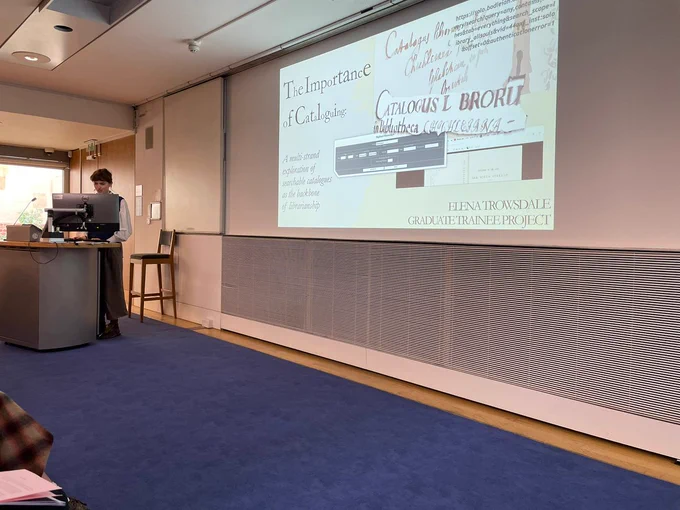 Elena, the trainee at All Souls is standing behind a computer. Her presentation, entitled 'The Importance of Cataloguing' is projected onto the screen. 