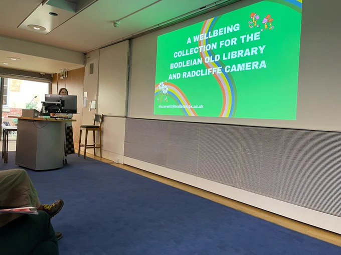 Nia, the trainee at the Old bod is standing behind a computer on the left of the image. On the right of the image, we see the first slide of her presentation, entitled ''A wellbeing collection for the Bodleian Old Library and Radcliffe Camera'. 