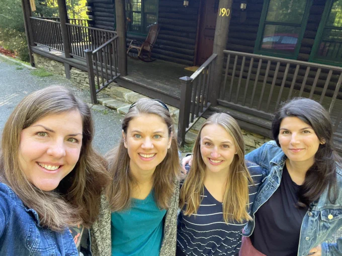 Photo of four women smiling and looking up at the camera. They are outside in front of a house and it is daytime.