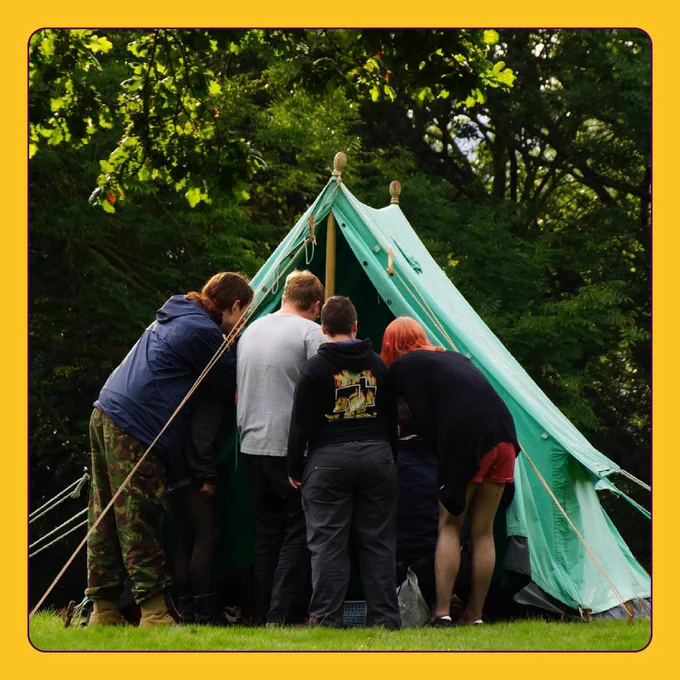 A yellow border around a square tile. In the centre is a photo of five people peering into a tent, surrounded by greenery.