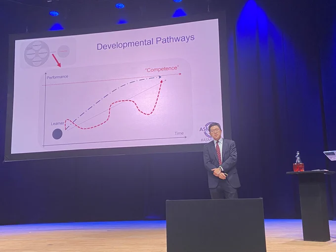 Photograph of prof yoon soo park on a large stage; hands clasped in front and speaking: large screen to the left with the title ‘developmental pathway’. There is a graph with a linear line; and a wiggly line