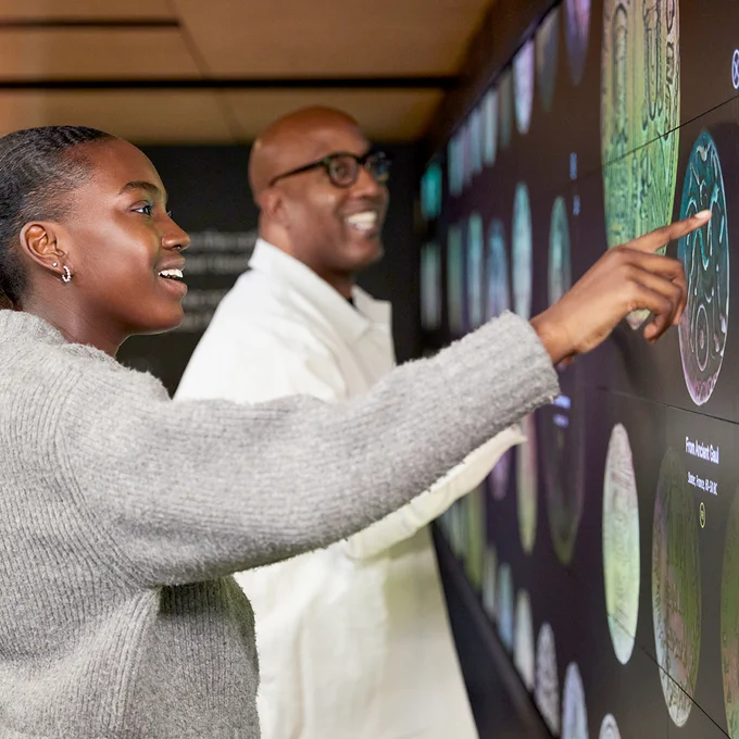 A smiling man and teen-aged girl in front of a wall-sized wall video monitor that they are touching, manipulating images of coins.