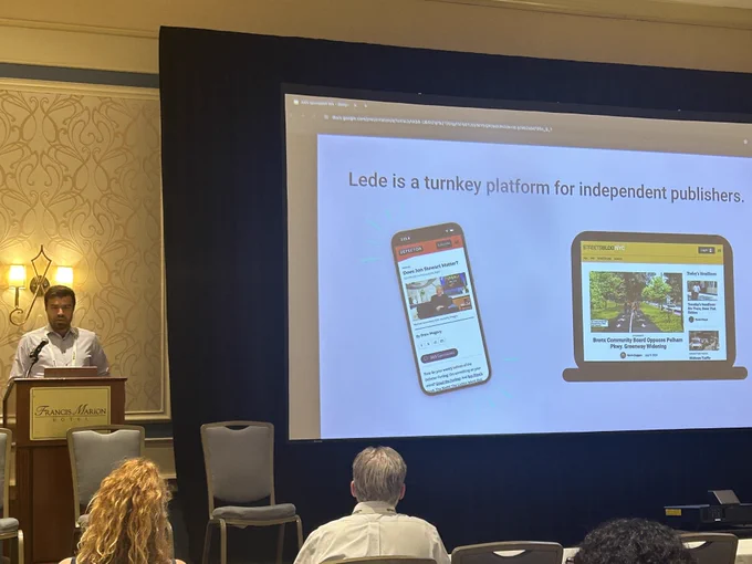 Lede CEO Matt Johnson standing at a lecturn in a hotel ballroom talking about Lede. To his right is a large screen with the heading "Lede is a turnkey platform for independent publishers" and two images. The image on the left is a mockup of a mobile device featuring the homepage of Defector, a Lede publisher. The other image is a mockup of a laptop featuring the home page of Streetsblog, another Lede publisher. Only the first row of people in the ballroom is visible.
