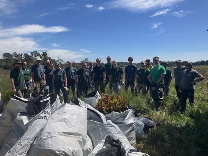 This image shows a group of volunteers. They are standing in Wareham Forest in front of large bags containing plants they have removed from the ground. 