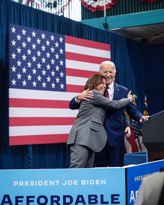 Vice President Kamala Harris and President Joe Biden hug on stage. Behind them is the American flag. 