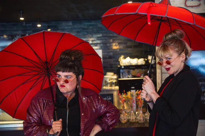 Two people holding red umbrellas indoors at a counter, with one person making a playful face and gesture while wearing heart-shaped sunglasses. The background suggests a casual, lively indoor environment, possibly a café or bar.