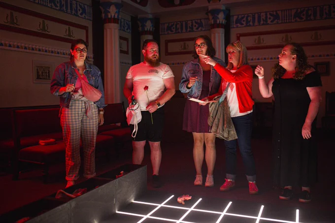A group of five people stand in a dimly lit theater, appearing engaged in a discussion or rehearsal, with one person pointing towards the stage that features glowing LED lights on the floor.