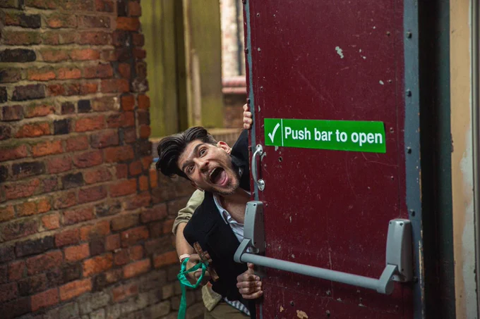 Person peering around a partially opened red door with a playful expression, holding the door handle. A sign on the door reads "Push bar to open." The setting has a backdrop of brick walls, suggesting an urban environment.