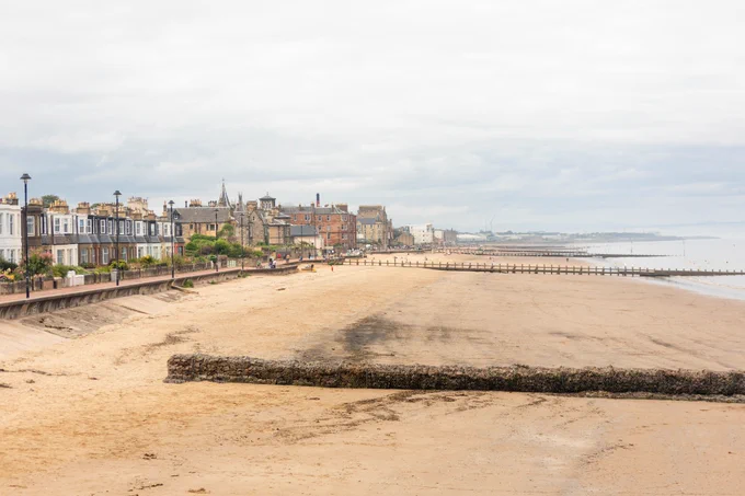 Colour photo of Portobello Beach.  The image shows a long beach with yellow sand, with houses overlooking the beach. The sky is cloudy with dark blue patches. Towards the right of the image, you can see a gentle sea with a few waves lapping the shore.
Photo credit: tekinturkdogan