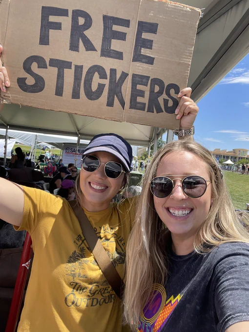 Two people smiling while holding a cardboard sign that says "FREE STICKERS" at an outdoor festival, with tents and people in the background.