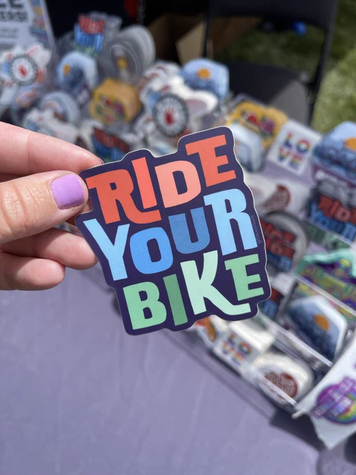A hand holding a colorful sticker with the text "Ride Your Bike," in front of a table covered with various stickers.