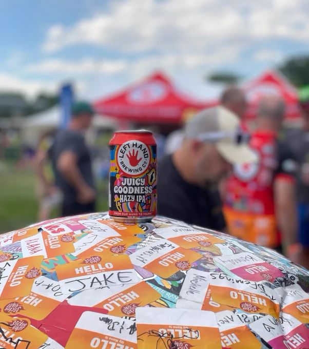A can of "Left Hand Brewing Co. Juicy Goodness Hazy IPA" sits on Saul the Sticker Ball with festival stickers, with people and event tents blurred in the background.